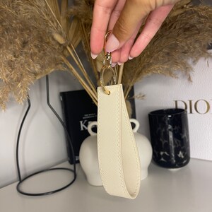 May include: A white leather keychain with a gold metal ring and clasp. The keychain has a textured surface and is a simple, elegant design.