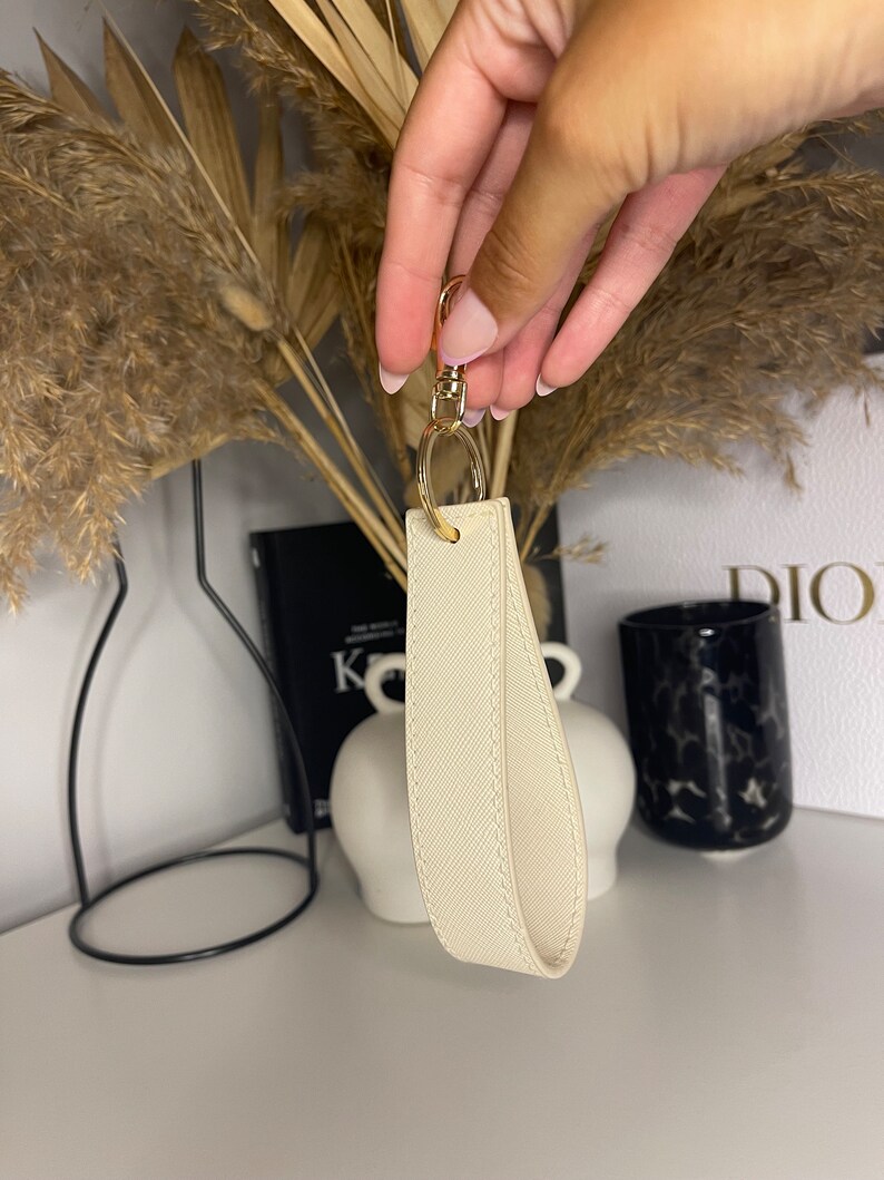 May include: A white leather keychain with a gold metal ring and clasp. The keychain has a textured surface and is a simple, elegant design.