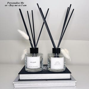 May include: Two glass diffuser bottles with black lids and black reeds. The bottle on the left has a label that says "UNTIL NAMED" and "LIME, BASIL AND MADAN". The bottle on the right has a label that says "MERRY CHRISTMAS MUM, LOVE FROM OLIVIA".