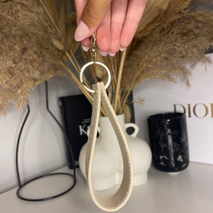May include: A cream-colored leather keychain with a gold metal ring. The keychain is shaped like a teardrop and has a smooth, polished finish.