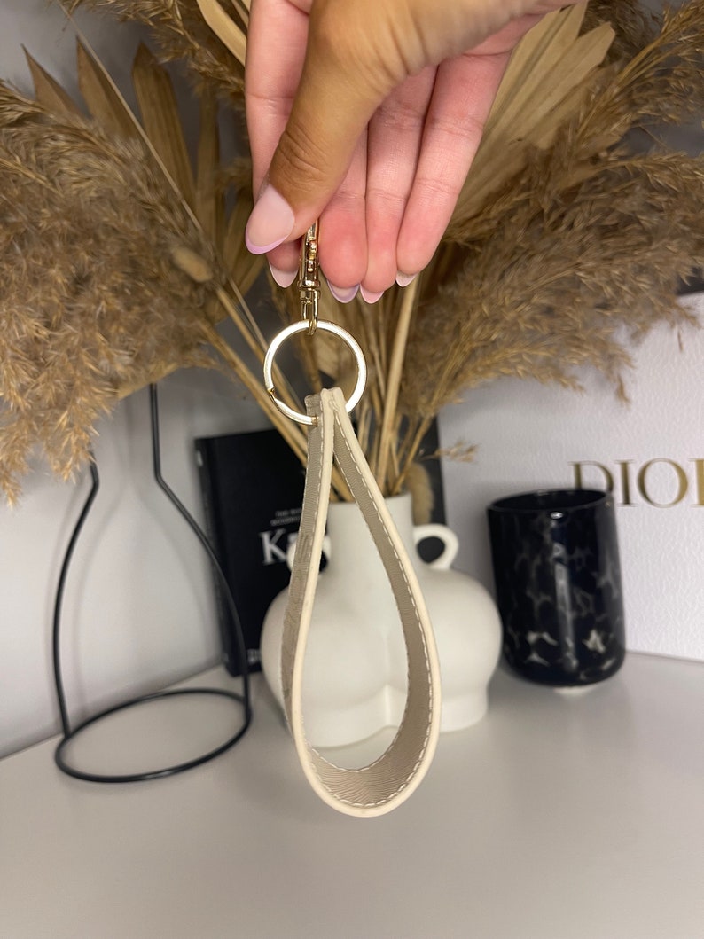 May include: A cream-colored leather keychain with a gold metal ring. The keychain is shaped like a teardrop and has a smooth, polished finish.