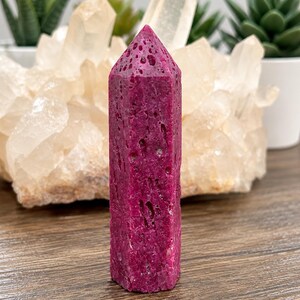 May include: A vibrant magenta, hexagonal crystal tower with a porous, textured surface. The stone features a pointed top and a deep, saturated pinkish-red color throughout its vertical, column-like structure.