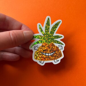 Könnte beinhalten: Ein glitzernder Ananas-Aufkleber mit einem lächelnden Gesicht, Armen und Beinen. Die Ananas hat einen strukturierten, orangefarbenen Körper und grüne Blätter. Der Aufkleber wird vor einem orangefarbenen Hintergrund gehalten.