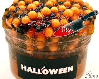 May include: A clear plastic container filled with orange and black slime, topped with orange and black beads. A small knife with a blood-splatter design is inserted into the slime. The container has a black label with the word "HALLOWEEN" in white.