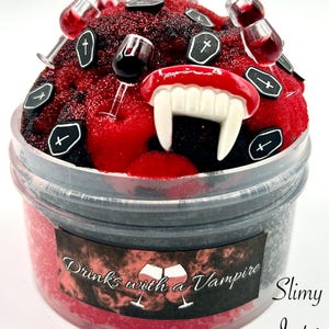 May include: A red and black slime with a vampire theme. The slime is in a clear plastic container with a label that says "Drinks with a Vampire". The slime is decorated with small plastic wine glasses, fangs, and cross-shaped charms.