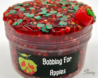 May include: A clear plastic container filled with red slime. The slime is decorated with green and red apple-shaped sprinkles and a red apple charm. The container has a black label that reads "Bobbing For Apples" with a green apple graphic.
