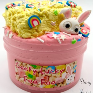 May include: A clear plastic container filled with pink slime, topped with yellow textured slime, and decorated with rainbow and star-shaped embellishments. A white bunny figure is on top. The container has a label that reads "Goofy Lucky Foo Foo".