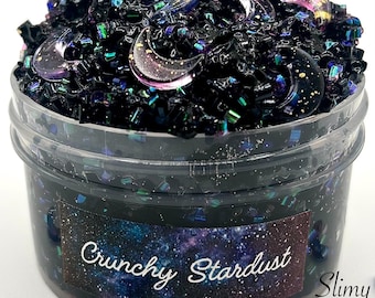 May include: Black slime with iridescent glitter and purple crescent moon charms in a clear plastic container. The label reads "Crunchy Stardust" with a galaxy background and "Slimy Jester" in the corner.