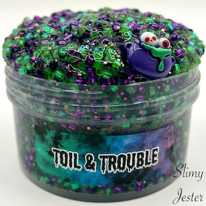 Puede incluir: Un tarro de plástico transparente lleno de slime verde y morado con purpurina negra y verde. El slime tiene un amuleto de cara de monstruo morado y verde en la parte superior. El tarro tiene una etiqueta que dice "Toil & Trouble" y "Slimy Jester".
