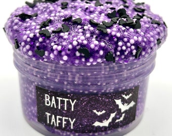 Könnte beinhalten: Lilafarbener Schleim mit weißen und schwarzen Flecken in Form von Fledermäusen in einem durchsichtigen Plastikbehälter mit einem Etikett, auf dem "Batty Taffy" steht.
