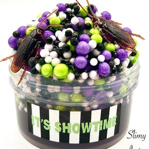 May include: A clear plastic container filled with slime, featuring purple, green, white, and black beads. Two brown cockroaches are partially submerged in the slime. The container has a black and white striped label that reads "IT'S SHOWTIME" and a smaller label that reads "Slimy Jester".