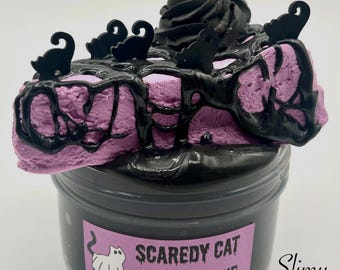 May include: A black container of slime with a purple and black cheesecake-shaped slime inside. The cheesecake is decorated with black cats and a black chocolate topping. The container has a label that reads "Scaredy Cat Cheesecake".