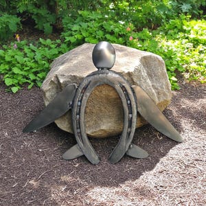 May include: A metal sculpture of an angel made from a horseshoe with wings and a round head. The sculpture is sitting on a large rock.