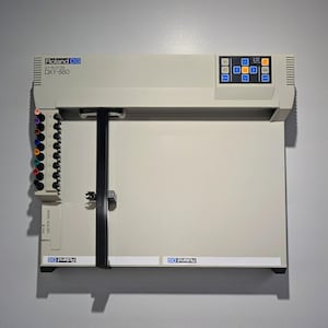 May include: A cream-colored Roland DXY-880 plotter machine with a control panel and a paper holder. The machine has a black strap and a series of colored pens. The Roland DG logo is visible.