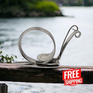 May include: A silver snail sculpture made from a bent spoon. The snail is sitting on a wooden surface with a blurry background of a lake and trees.