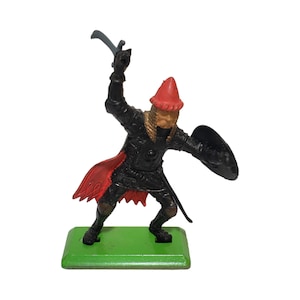 May include: A black plastic toy knight figure with a red and black cape, holding a silver sword and a black shield. The figure is standing on a green base.