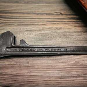 Puede incluir: Una llave de tubo negra con el texto "TRADE RIDGID MARK PAT. 1727823 1B" en el mango.