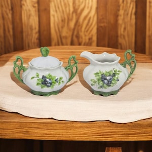 May include: A white porcelain sugar bowl and creamer set with green handles and a blueberry design. The set is on a white tablecloth.