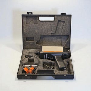 Puede incluir: Un dispositivo electrónico negro y gris con un asa, en un estuche negro abierto. El estuche contiene unos auriculares con orejeras naranjas, una pequeña varilla plateada y otros accesorios. El dispositivo tiene un panel plateado y naranja.