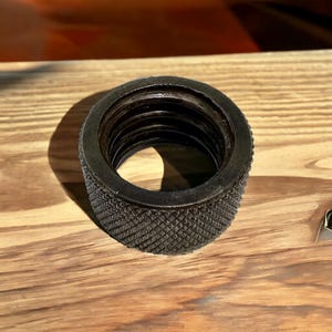 Puede incluir: Un anillo de metal negro con una superficie exterior texturizada y una superficie interior lisa. El anillo tiene una serie de ranuras conc&eacute;ntricas en el interior.