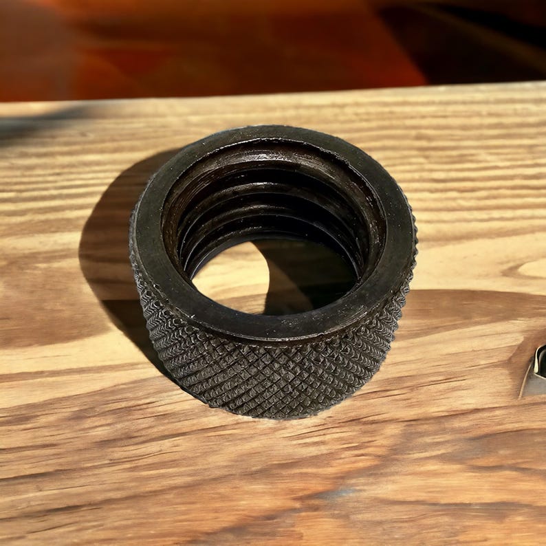 Puede incluir: Un anillo de metal negro con una superficie exterior texturizada y una superficie interior lisa. El anillo tiene una serie de ranuras conc&eacute;ntricas en el interior.