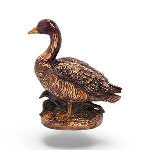 Könnte beinhalten: Eine Keramik-Gänsefigur mit braunen und goldenen bemalten Federn, die auf einem Sockel mit grünen Blättern steht. Die Gans hat einen langen Hals und einen kleinen Schnabel.