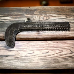Puede incluir: Una llave de tubo negra con la palabra "RIDGID" impresa en el mango. La llave tiene una mand&iacute;bula dentada y una escala marcada en pulgadas.