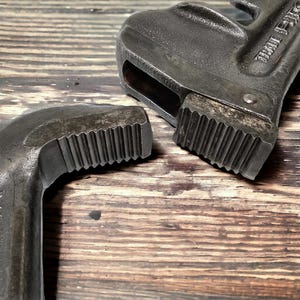Puede incluir: Primer plano de una llave de tubo negra con una mand&iacute;bula dentada. La llave es de metal y tiene una superficie texturizada. La llave est&aacute; sobre una superficie de madera.