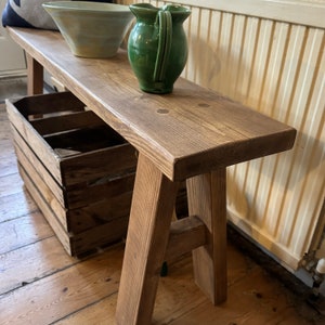 May include: A rustic wooden bench with a natural brown finish. The bench has a simple A-frame design and is made from reclaimed wood. It is sitting on a wooden floor and has a wooden crate underneath.