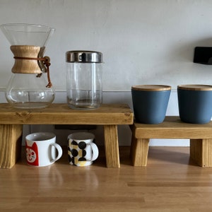 May include: Two wooden shelves with two mugs, a glass jar with a lid, and two blue ceramic containers. The mugs have a cat and a geometric pattern. The shelves are made of natural wood.