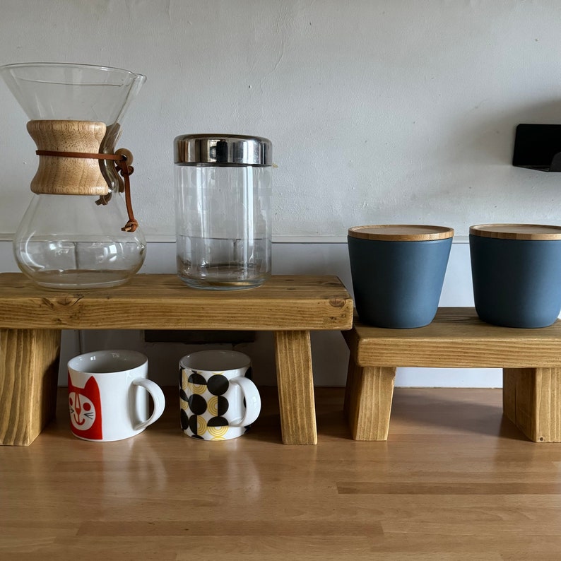 May include: Two wooden shelves with two mugs, a glass jar with a lid, and two blue ceramic containers. The mugs have a cat and a geometric pattern. The shelves are made of natural wood.
