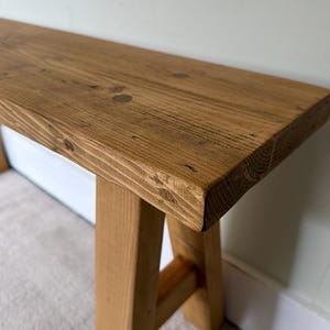 May include: A rustic wooden bench with a natural finish. The bench features a rectangular top and sturdy, angled legs. The wood grain is visible, showcasing the natural knots and textures of the material. The bench is a light brown color.