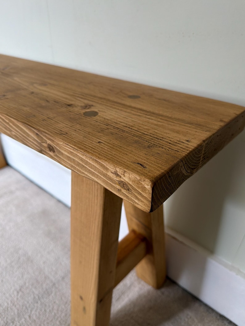 May include: A rustic wooden bench with a natural finish. The bench features a rectangular top and sturdy, angled legs. The wood grain is visible, showcasing the natural knots and textures of the material. The bench is a light brown color.
