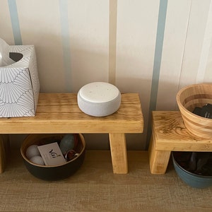 May include: Two wooden stools with a white tissue box, a grey smart speaker, and a wooden bowl on top. The stools have a black bowl and a blue bowl underneath.