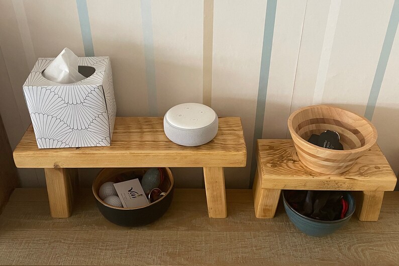 May include: Two wooden stools with a white tissue box, a grey smart speaker, and a wooden bowl on top. The stools have a black bowl and a blue bowl underneath.