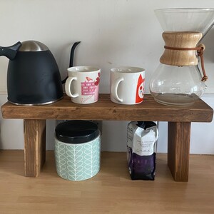 May include: A wooden shelf with two mugs, a black kettle, a glass coffee pot, a jar with a green and white pattern, and a bag of coffee beans. The shelf is brown and has a rustic look.