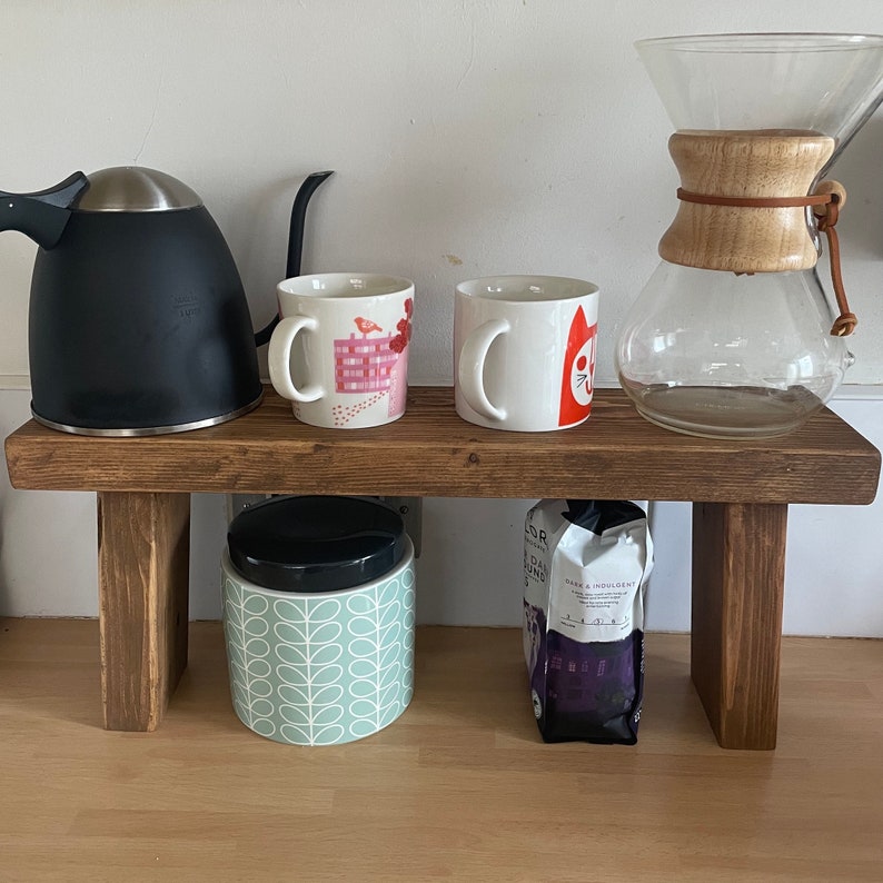 May include: A wooden shelf with two mugs, a black kettle, a glass coffee pot, a jar with a green and white pattern, and a bag of coffee beans. The shelf is brown and has a rustic look.