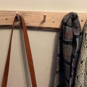 May include: A wooden wall-mounted coat rack with four hooks. A brown leather strap hangs from one hook, and two scarves hang from the other two hooks. One scarf is a plaid pattern in grey, black, and red, and the other is a cream-coloured crocheted scarf.