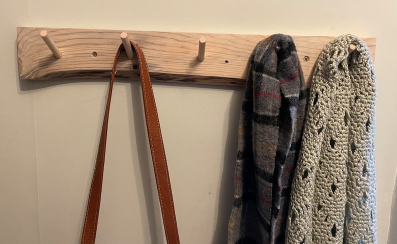 May include: A wooden wall-mounted coat rack with four hooks. A brown leather strap hangs from one hook, and two scarves hang from the other two hooks. One scarf is a plaid pattern in grey, black, and red, and the other is a cream-coloured crocheted scarf.