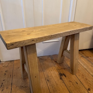 May include: A small, natural wood bench with a rectangular seat and A-frame legs. The bench is made of light-colored wood with visible grain and knots. The bench is approximately 36 inches long.