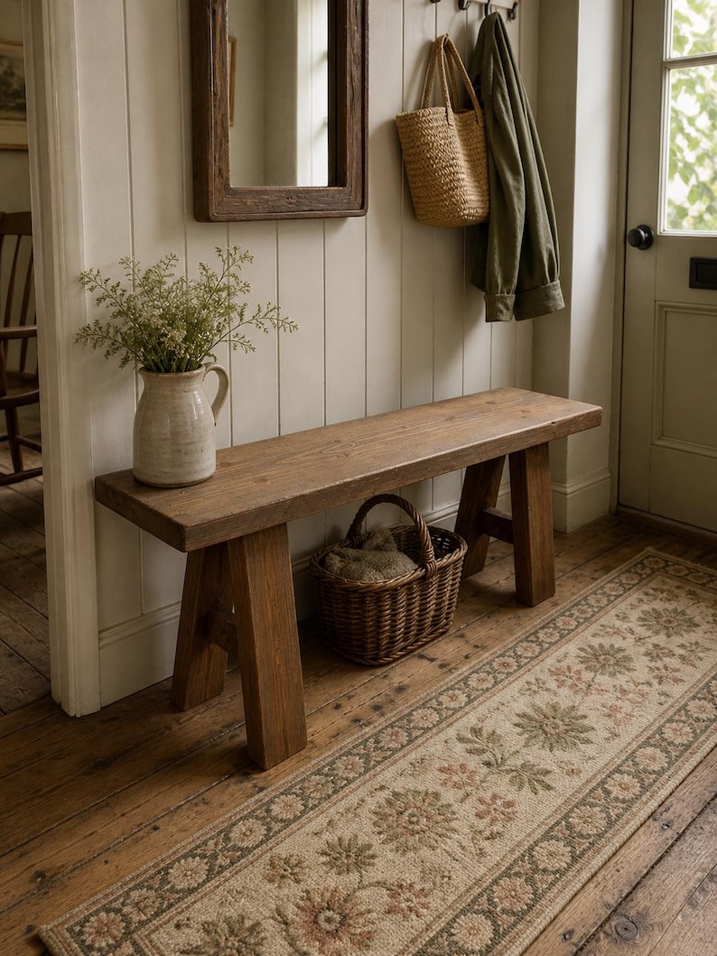May include: A wooden bench with a natural finish in a hallway setting. A woven basket is placed beneath the bench, and a floral runner rug leads towards a door. A vase with greenery and a mirror are also visible.