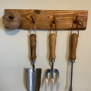 May include: A wooden wall-mounted rack with three hooks, holding a trowel, a hand fork, and a hand cultivator. The rack is stained a dark brown colour. A ball of twine is hanging on the left side of the rack.