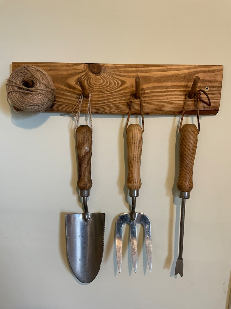 May include: A wooden wall-mounted rack with three hooks, holding a trowel, a hand fork, and a hand cultivator. The rack is stained a dark brown colour. A ball of twine is hanging on the left side of the rack.