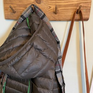 May include: A wooden coat rack with three hooks. A brown puffer jacket with a plaid scarf is hanging on one hook. Two brown leather straps are hanging on the other hooks.