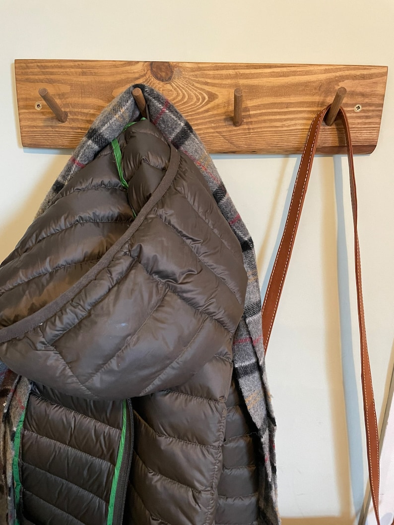 May include: A wooden coat rack with three hooks. A brown puffer jacket with a plaid scarf is hanging on one hook. Two brown leather straps are hanging on the other hooks.