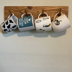 May include: Four white ceramic mugs with different designs hang on a wooden rack. The mugs feature black cats, penguins, and the text "Keep Calm and Feed the Ducks".