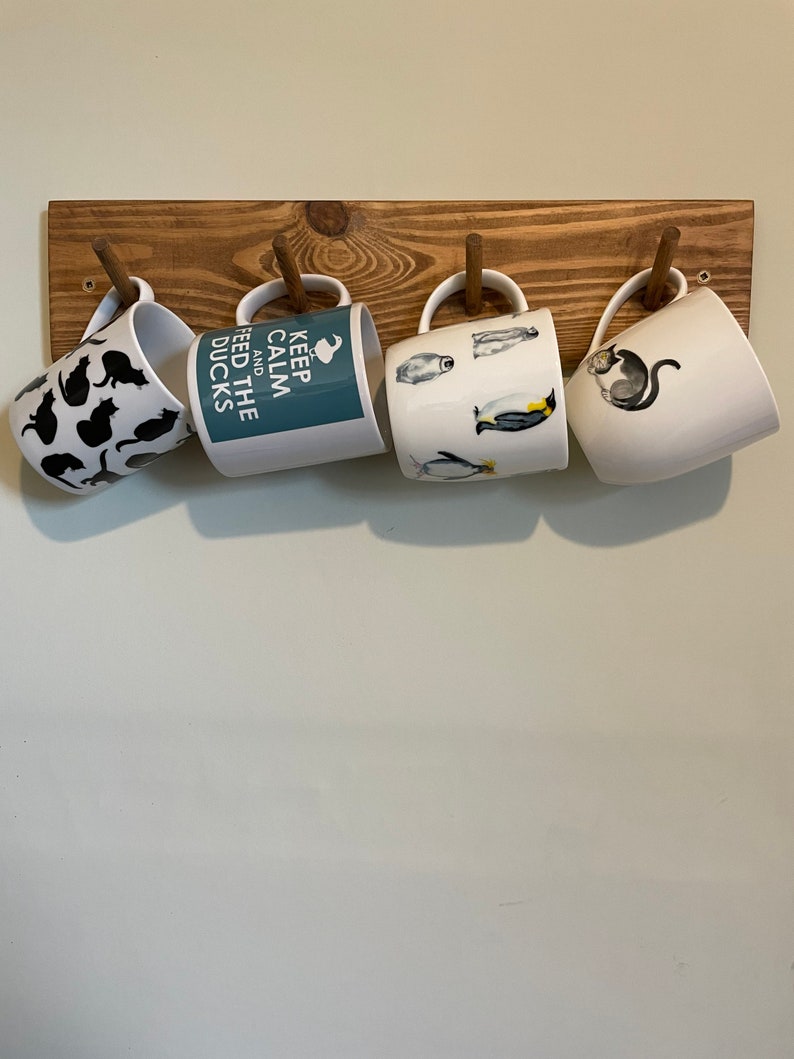 May include: Four white ceramic mugs with different designs hang on a wooden rack. The mugs feature black cats, penguins, and the text "Keep Calm and Feed the Ducks".