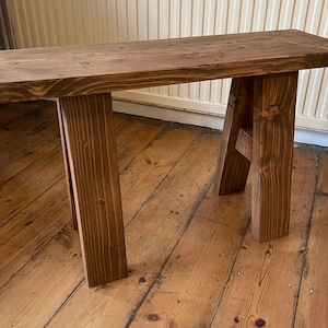 May include: A rustic wooden bench with a dark brown stain. The bench has a simple design with a single plank top and two A-frame legs.