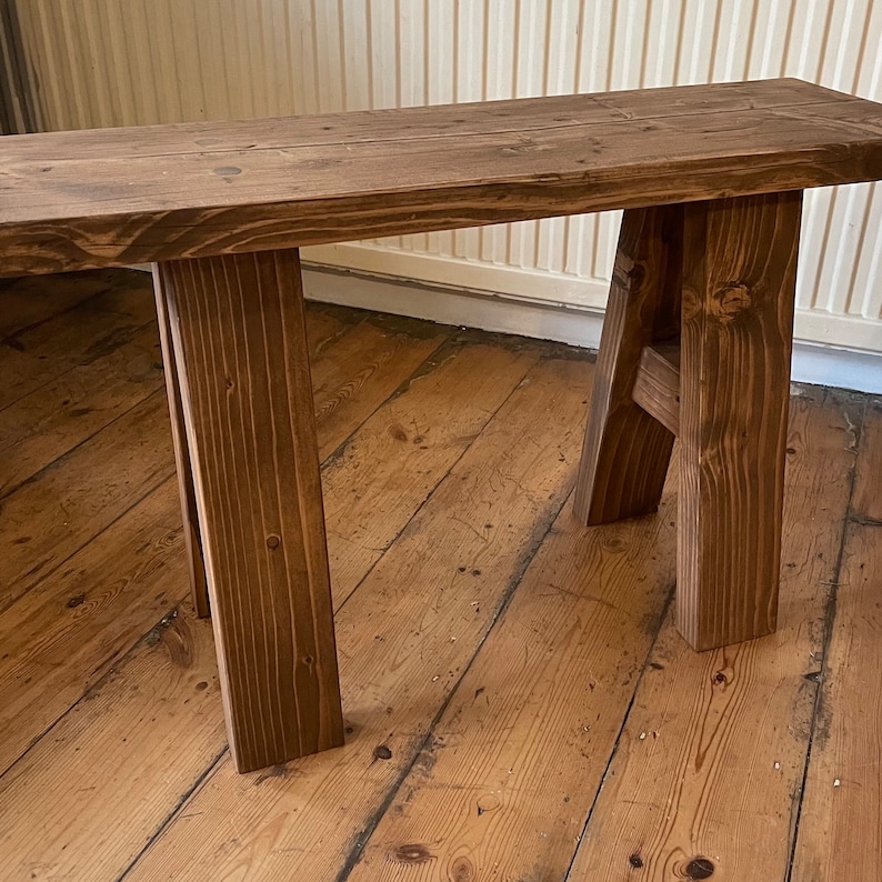 May include: A rustic wooden bench with a dark brown stain. The bench has a simple design with a single plank top and two A-frame legs.