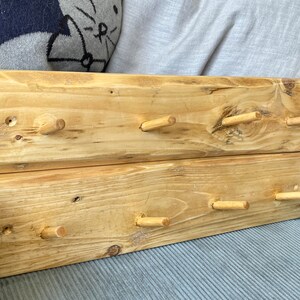May include: A wooden wall-mounted coat rack with six wooden pegs. The rack is made of two pieces of reclaimed wood, stained in a light brown colour.